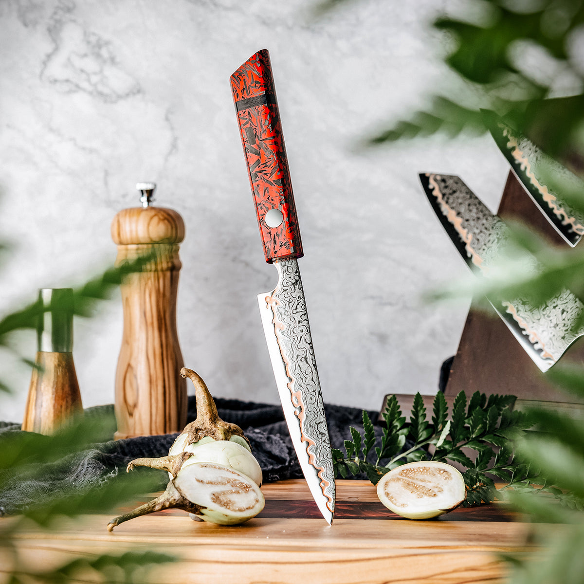 Cuchillo Utilitario 13cm 10Cr+Lluvia Cobre Damasco54 Resina Roja y Carbono apuntando tabla de madera con molidor de pimienta al fondo