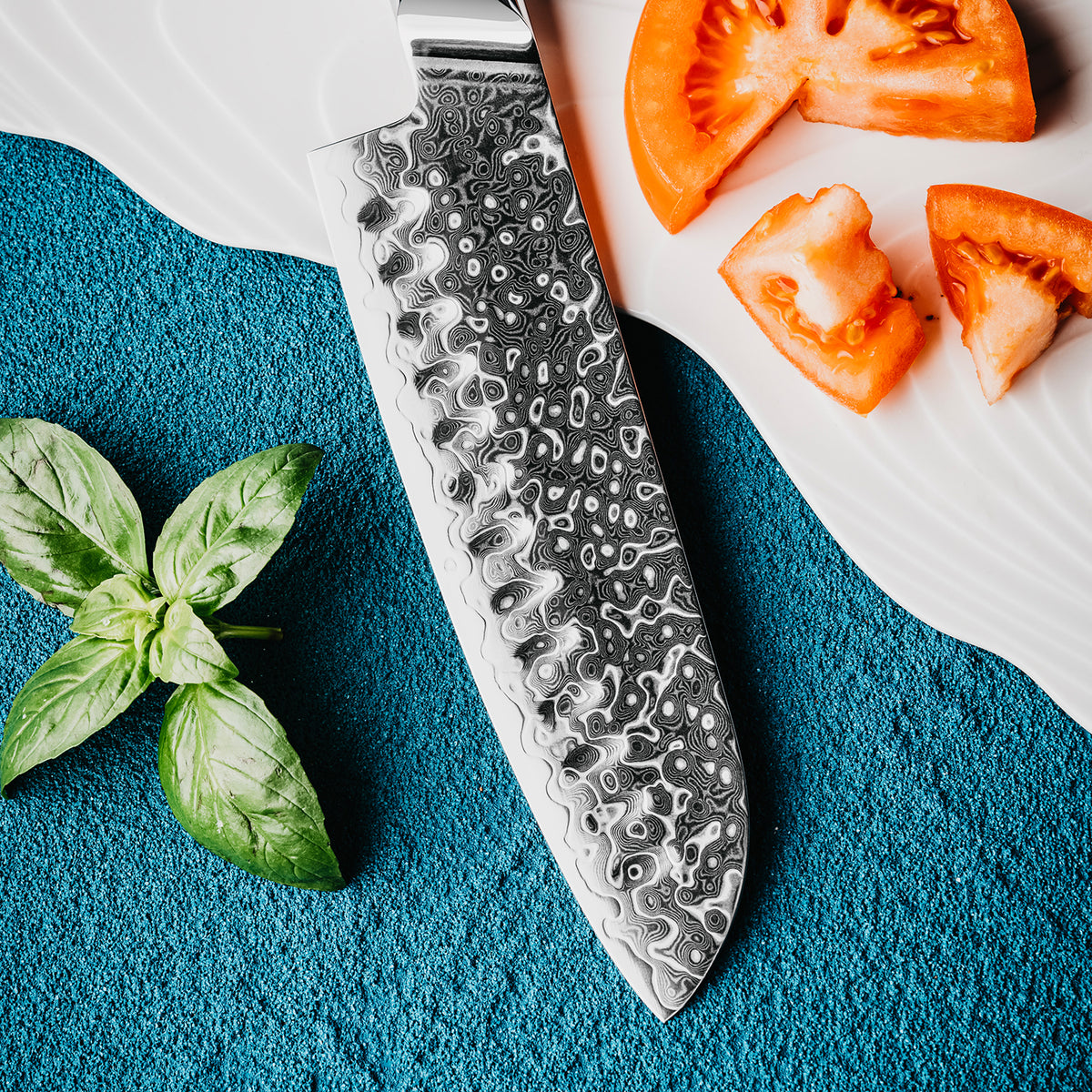 Cuchillo Santoku 18cm 10Cr+Gotas Damasco128 Full Tang Resina Azul, compañado de tomate y estrella de hojas de albaca.
