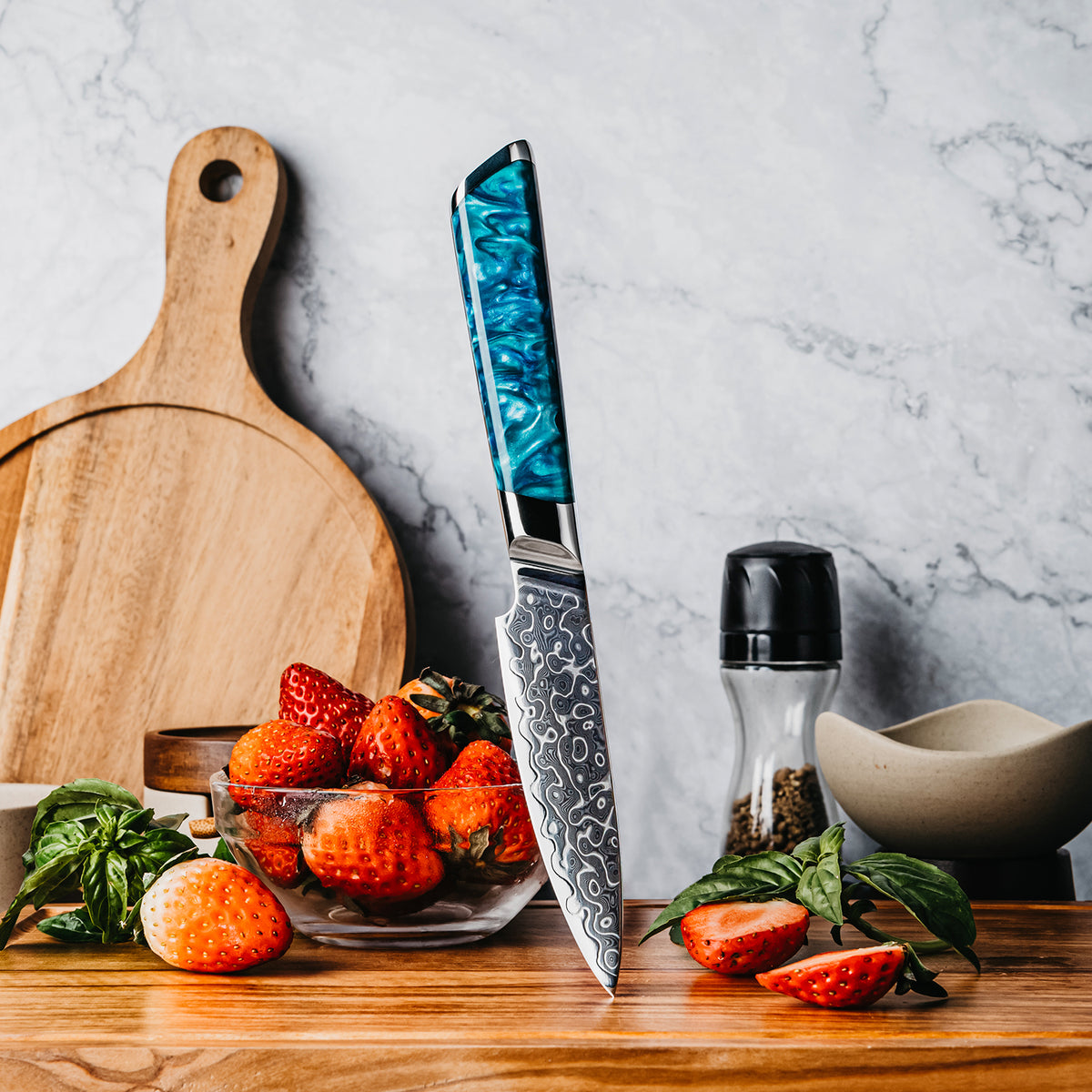 Cuchillo Puntilla 10cm 10Cr+Gotas Damasco128 Full Tang Resina Azul apuntando tabla de madera acompañado con fresas cortadas y molidor de pimienta