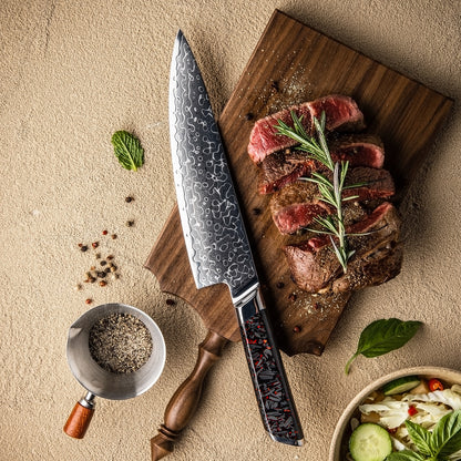 Cuchillo de Chef 20cm 10Cr+Gotas Damasco128 Full Tang Resina Carbono, sobre una tabla de picar, acompañado de un seak a tres cuartos con romero y pimienta.