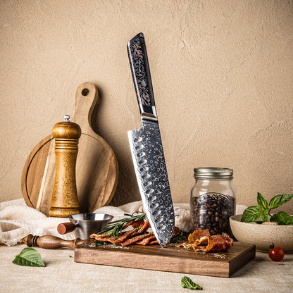 Cuchillo Santoku 18cm 10Cr+Gotas Damasco128 Full Tang Resina Carbono, apuntando una tabla de picar, acompañado de tocino y especias.