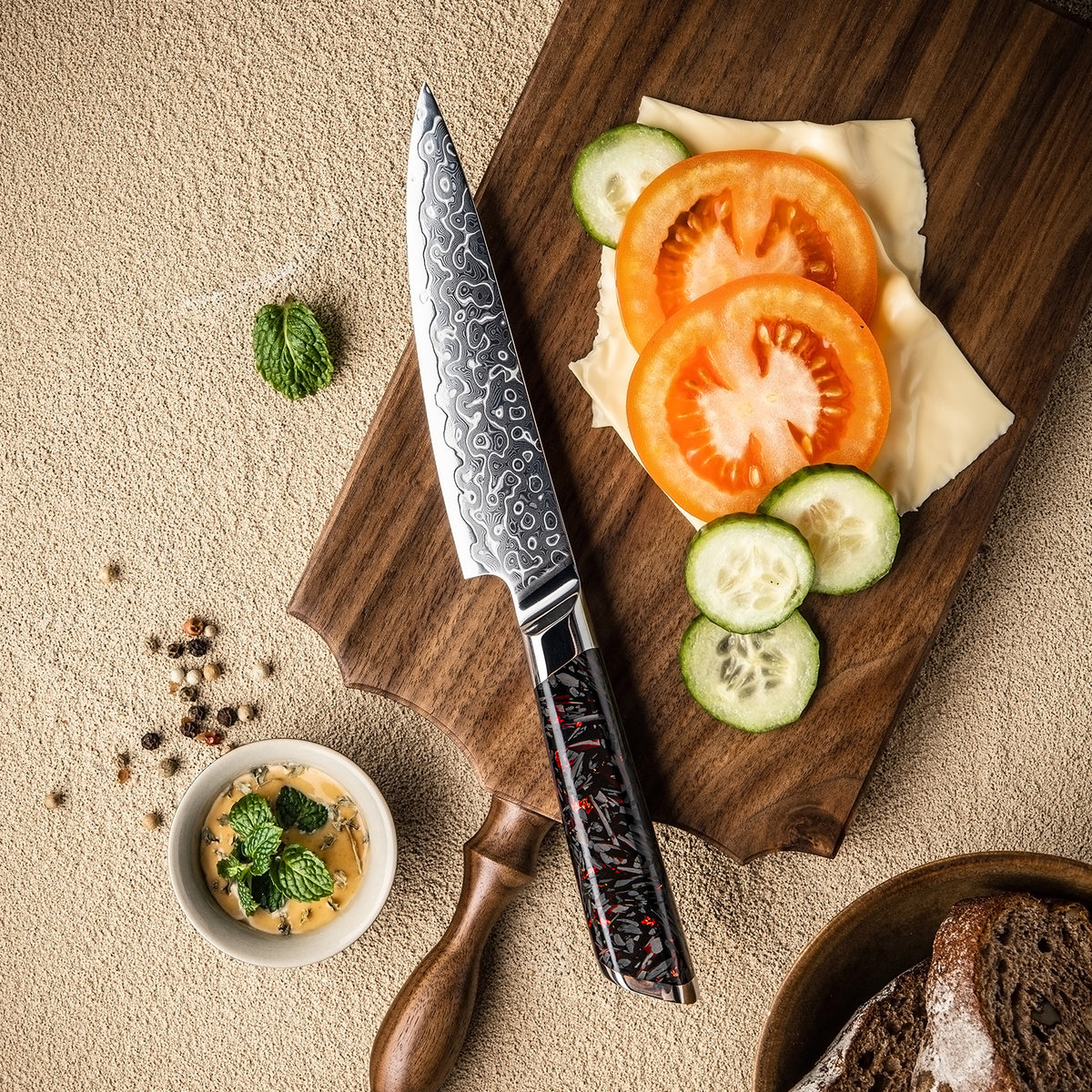 Cuchillo Utilitario 13cm 10Cr+Gotas Damasco128 Full Tang Resina Carbono, cuchillo en el centro, sobre una mesa de picar, verduras.