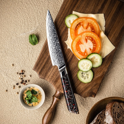 Cuchillo Utilitario 13cm 10Cr+Gotas Damasco128 Full Tang Resina Carbono, cuchillo en el centro, sobre una mesa de picar, verduras.