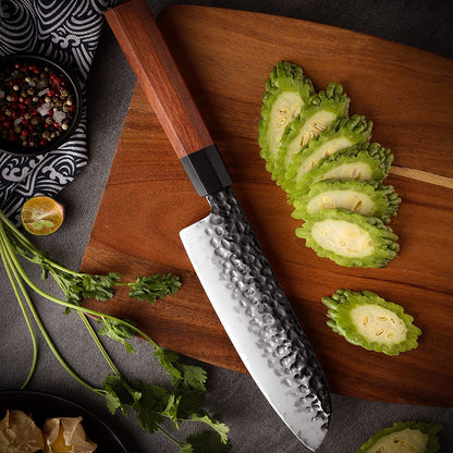 Cuchillo Santoku 19cm AUS-10 Clad Kurouchi Martillado Mango Mkuruti y Ebano, sobre tabla de picar, acompañado de verduras.