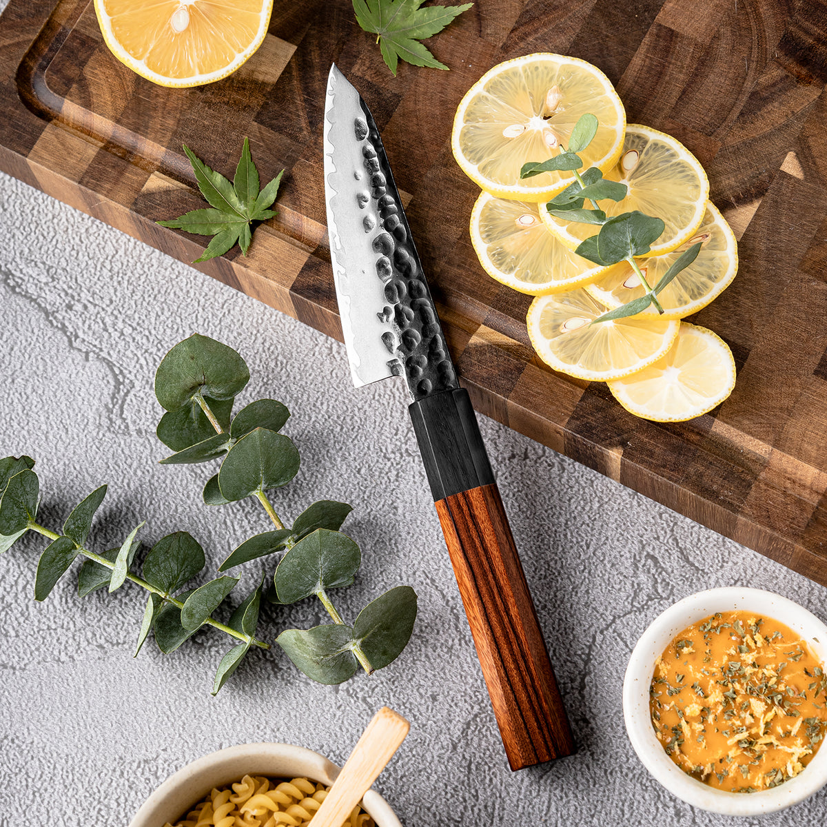 Cuchillo Puntilla 10cm AUS-10 Clad Kurouchi Martillado Mango Mkuruti y Ebano, sobre tabla de picar, acompañado de limones.